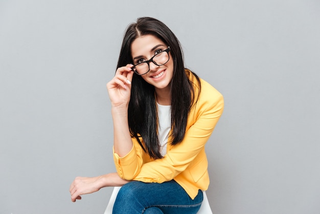 Felice giovane donna che indossa gli occhiali e vestita con una giacca gialla seduta su uno sgabello su una superficie grigia mentre tocca gli occhiali e guarda in avanti.