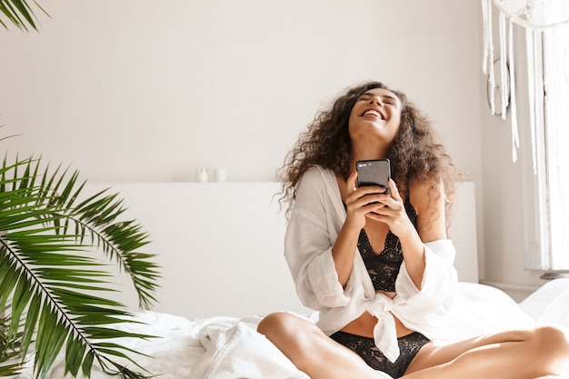 felice giovane donna che indossa lingerie sorridente e tenendo lo smartphone sul letto in appartamento bianco