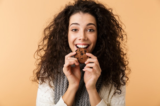 Felice giovane donna che indossa sciarpa invernale in piedi isolato sul muro beige, mangiando biscotti al cioccolato