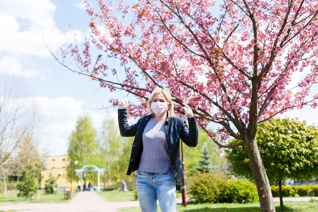 Felice giovane donna che indossa una maschera facciale per la protezione dai virus in piedi all'aperto nella soleggiata giornata primaverile.