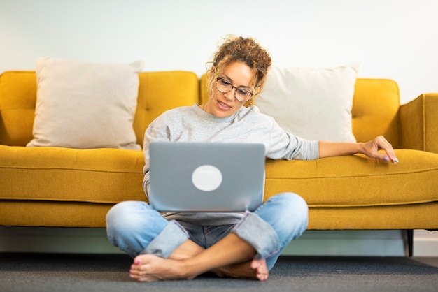 Felice giovane donna che lavora al computer portatile seduto sul pavimento contro il divano nel soggiorno di casa. Imprenditrice in casual che lavora da casa. Donna caucasica in occhiali utilizzando laptop.