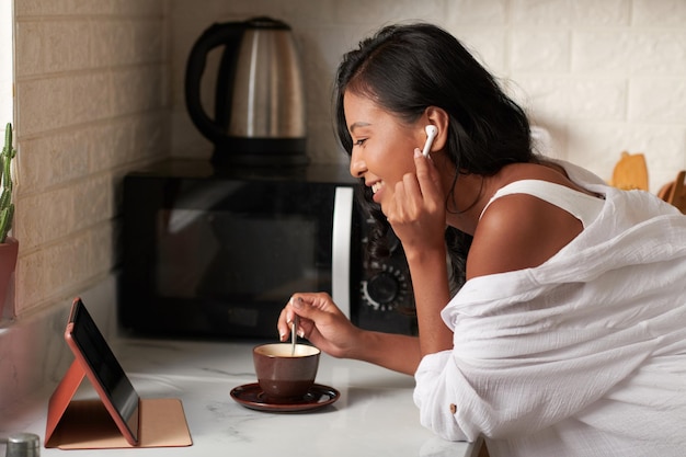 Felice giovane donna che mescola lo zucchero nel suo caffè quando partecipa al briefing online al mattino