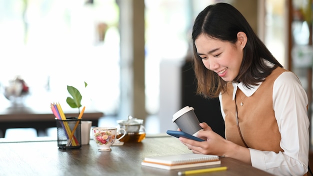 Felice giovane donna che utilizza smartphone navigando sui social media e bevendo caffè in ufficio.