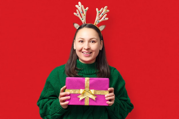 Felice giovane donna con corna di cervo di Natale con regalo su sfondo rosso