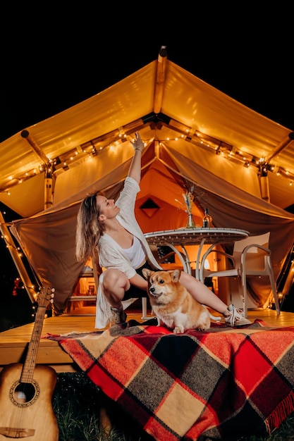 Felice giovane donna con il suo cane Welsh Corgi Pembroke che si rilassa nel glamping la sera d'estate vicino al falò accogliente Tenda da campeggio di lusso per attività ricreative all'aperto e attività ricreative Concetto di stile di vita