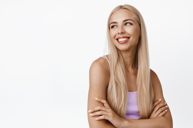 Felice giovane donna con lunghi capelli biondi e corpo in forma incrociare le braccia sul petto guardando dietro la spalla con sfondo bianco sorriso spensierato