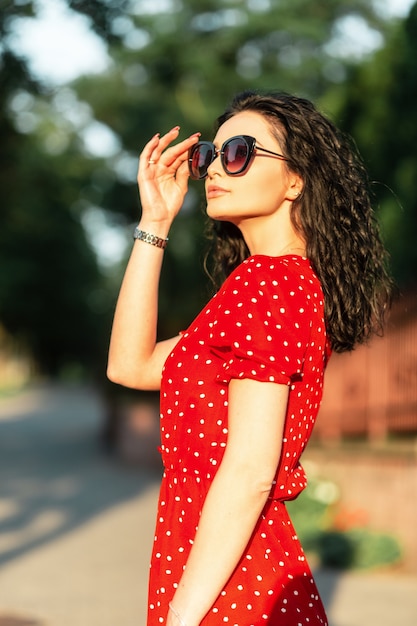 Felice giovane donna con occhiali da sole alla moda in abito rosso estivo vintage cammina in città al tramonto