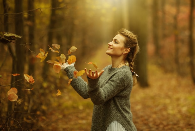 Felice giovane donna con trecce intrecciate vomita foglie cadute nelle sue mani nella foresta autunnale. Ritratto d'arte autunnale