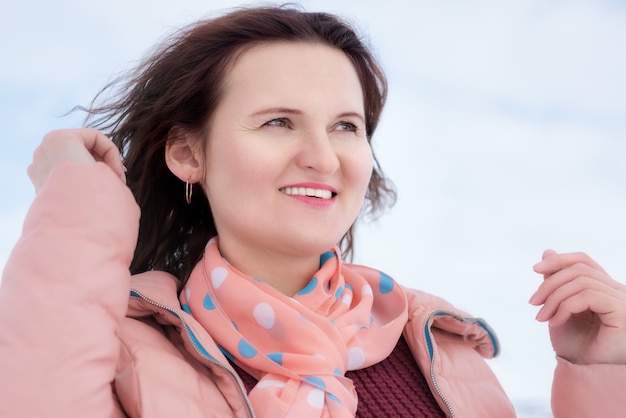 Felice giovane donna con un bel sorriso e capelli castani ricci che fluttuano nel vento, vestita con giacca beige, sciarpa intorno al collo. Ritratto di belle donne all'aperto in inverno.