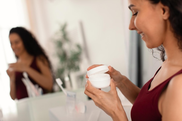 Felice giovane donna con un vasetto di crema per il viso in piedi davanti allo specchio applicando una crema idratante a casa