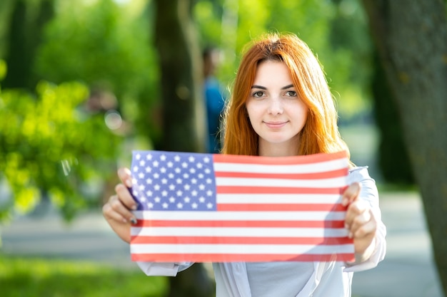 Felice giovane donna dai capelli rossi che tiene la bandiera nazionale degli Stati Uniti nelle sue mani all'aperto in estate. Ragazza positiva che celebra il giorno dell'indipendenza degli Stati Uniti.