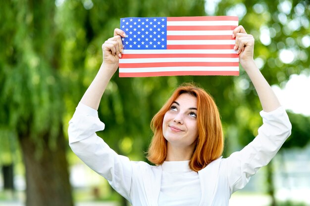 Felice giovane donna dai capelli rossi in posa con la bandiera nazionale USA sopra la sua testa all'aperto nel parco estivo