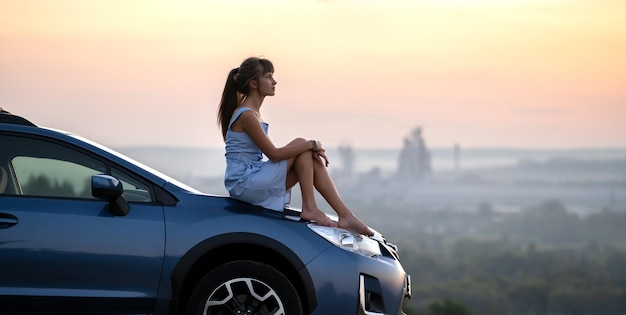 Felice giovane donna driver in abito blu godendo di una calda serata estiva seduta sul cofano della sua auto Concetto di viaggio e vacanza