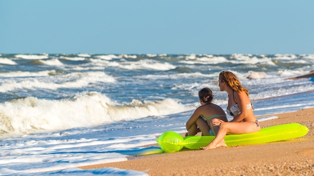 Felice giovane donna e sua figlia sono seduti su un materasso ad aria verde sulla riva e aspettano un'onda del mare in tempesta in una soleggiata giornata estiva durante le vacanze. Concetto di viaggio e tempo libero.