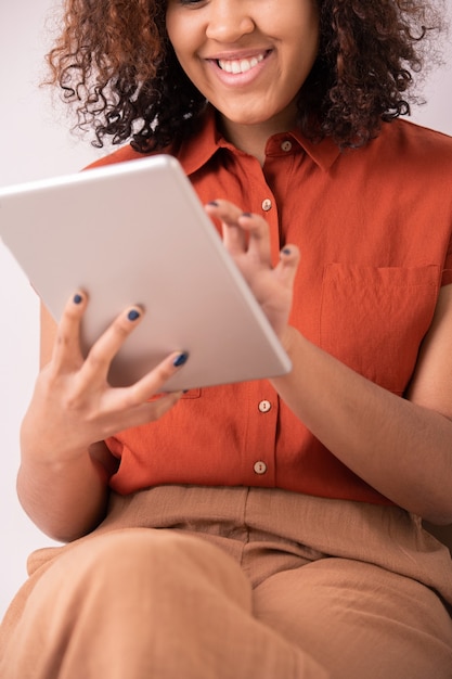 Felice giovane donna elegante con capelli ricci e sorriso a trentadue denti toccando lo schermo del tablet durante lo scorrimento dei post nei social network