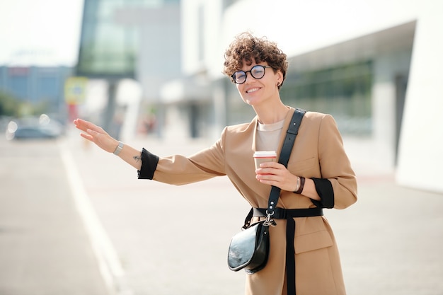 Felice giovane donna elegante con un bicchiere di caffè in piedi per strada e prendere un taxi contro un edificio moderno in ambiente urbano