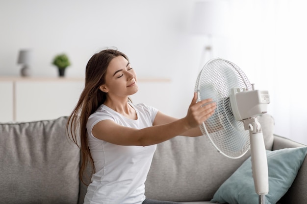 Felice giovane donna europea soffre di calore si siede sul divano prende l'aria fredda gira il ventilatore a se stessa