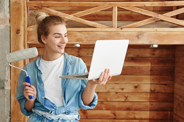 Felice giovane donna in abiti da costruzione con piani per laptop e fa riparazioni sullo sfondo del letto a castello in legno fatto in casa, concetto fai-da-te