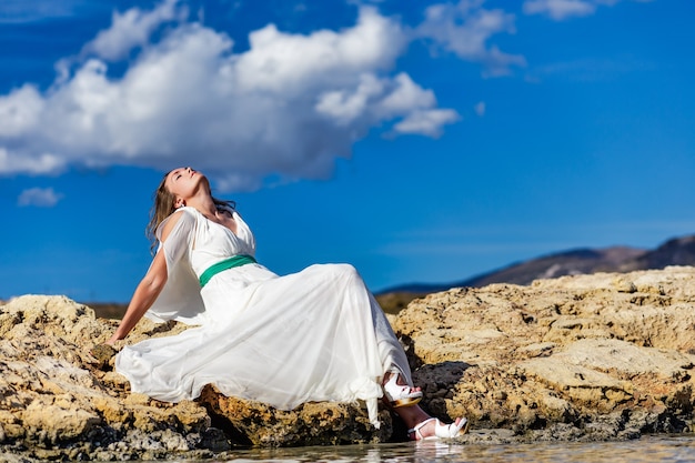 Felice giovane donna in abito bianco seduta sulla roccia di sabbia e godendo sulla spiaggia di elafonisi