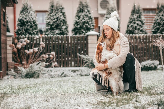 Felice giovane donna in bianco cappello lavorato a maglia cammina un cane nel giardino innevato. Cavalier King Charles Spaniel. Paesaggio invernale. Animali domestici. Momenti meravigliosi della vita. Molta felicità. Amore e cura.