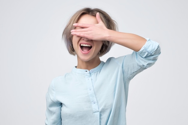 Felice giovane donna in camicia blu che copre i suoi occhi su sfondo grigio