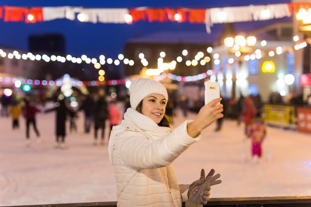 Felice giovane donna in inverno sulla pista di pattinaggio di scattare una foto su smartphone, selfie.