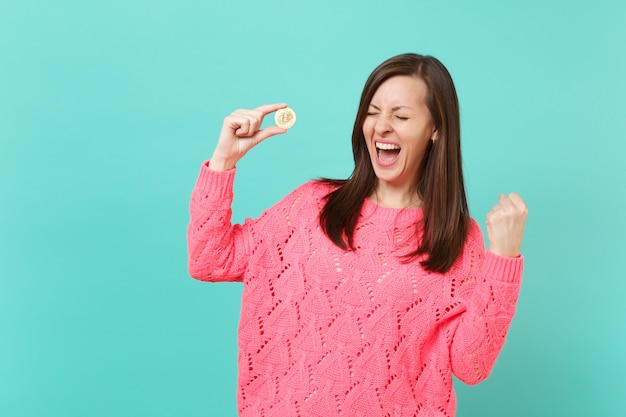 Felice giovane donna in maglia maglione rosa urlando facendo gesto vincitore tenere in mano bitcoin, valuta futura isolata su sfondo blu muro turchese. Concetto di stile di vita della gente. Mock up copia spazio.