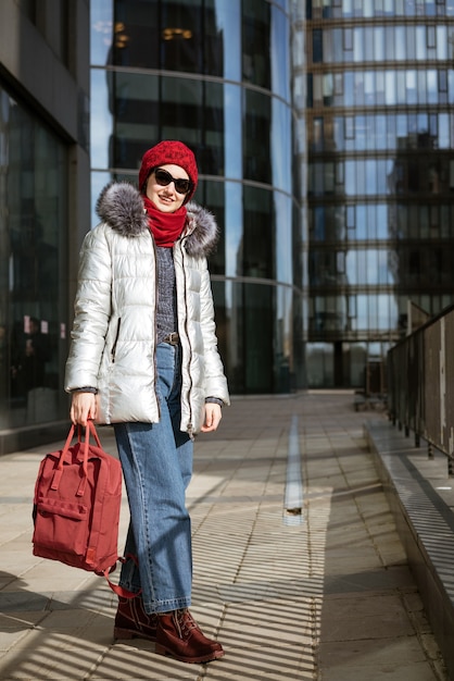 Felice giovane donna in occhiali da sole in una giacca invernale con uno zaino vicino all'edificio. Concetto di viaggio