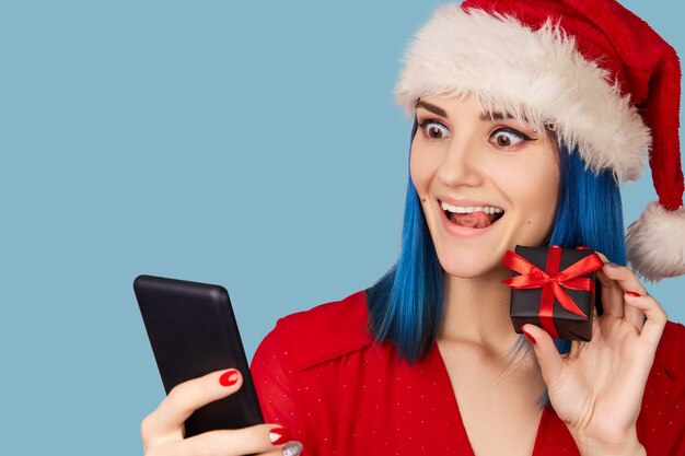 Felice giovane donna in santa hat con confezione regalo e smartphone su sfondo blu