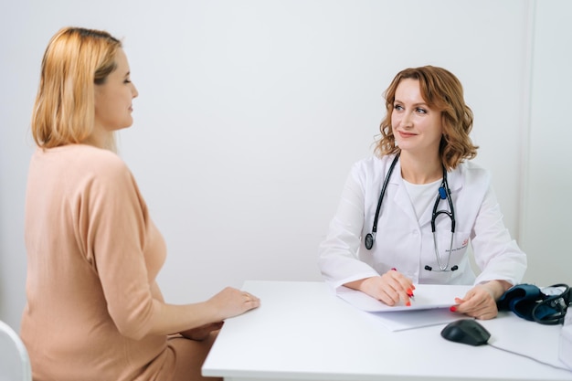 Felice giovane donna incinta con capelli biondi visita medico in clinica prenatale. Medico d'ascolto della bella donna incinta che si siede allo scrittorio. Concetto di gravidanza felice e sana.