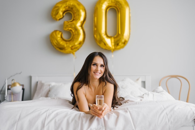 Felice giovane donna incontra il suo trentesimo compleanno a letto con in mano un bicchiere di champagne e sorridente