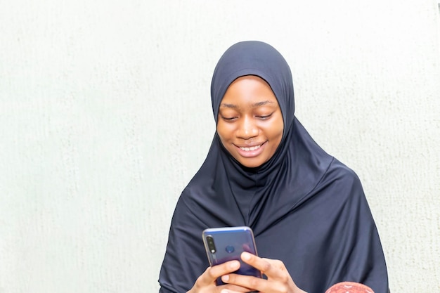 Felice giovane donna musulmana nigeriana vestita di hijab u tiene il telefono cellulare scarica la musica per la playlist ha un'espressione felice isolata su sfondo bianco