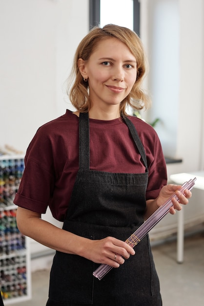 Felice giovane donna professionista lampworker in grembiule che tiene mazzetto di lunghi pezzi di vetro legati con filo mentre si trova in officina