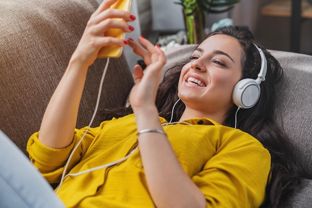 Felice giovane donna sdraiata sul divano con smartphone e cuffie per ascoltare musica a casa