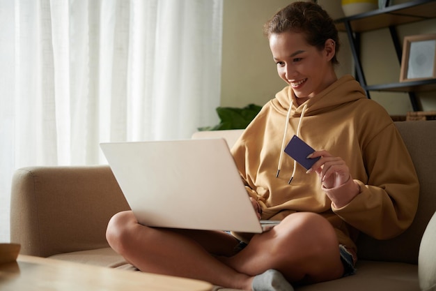 Felice giovane donna seduta sul divano con il computer portatile e utilizzando la carta di credito che fa shopping online a casa