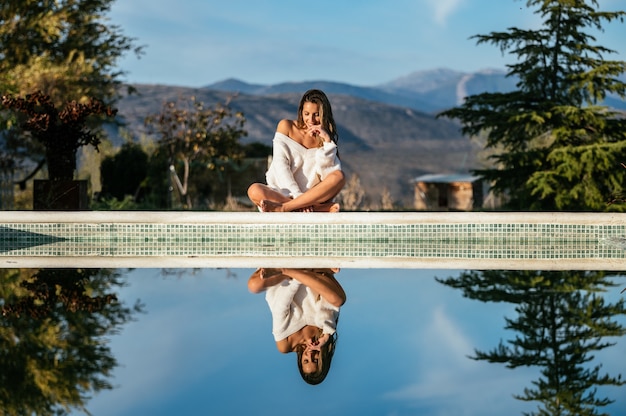 Felice giovane donna sorridente e toccando il viso mentre è seduto a gambe incrociate sul bordo della piscina e guardando l'acqua calma in cortile