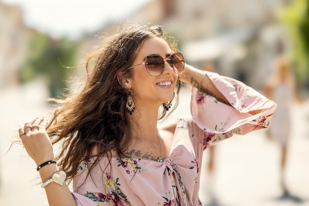 Felice giovane donna sorridente in abito estivo con occhiali da sole che cammina in città con il vento che le soffia i capelli