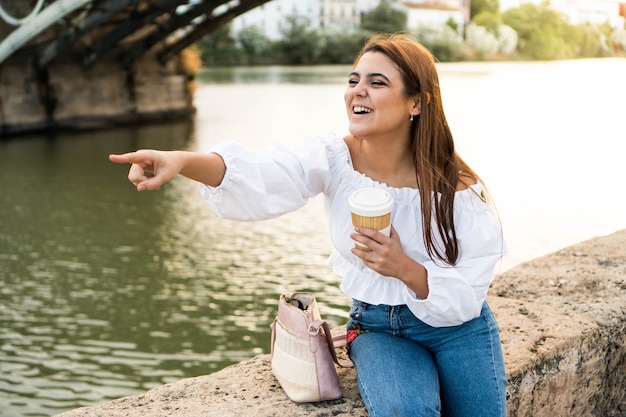 Felice giovane donna sorridente indicando i panorami e bevendo caffè in riva al fiume
