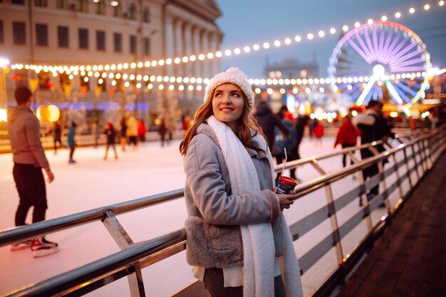 Felice giovane donna sul mercatino di Natale con caffè vicino alla pista di pattinaggio. Luci intorno. Vacanze invernali.