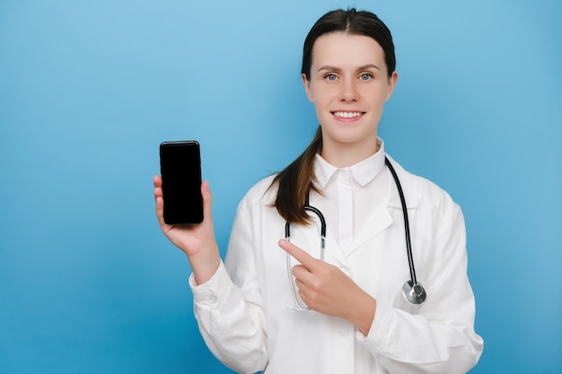 Felice giovane dottoressa che punta il dito sullo schermo del cellulare, consiglia il controllo del download, l'applicazione di consultazione, isolata su sfondo blu. Covid-19, operatori sanitari e concetto di medicina online