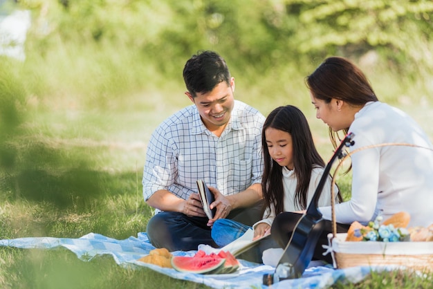 Felice giovane famiglia asiatica padre, madre e bambino bambina che si diverte e si diverte all'aperto sulla coperta da picnic leggendo il libro nel parco al sole, concetto di primavera per il tempo libero estivo