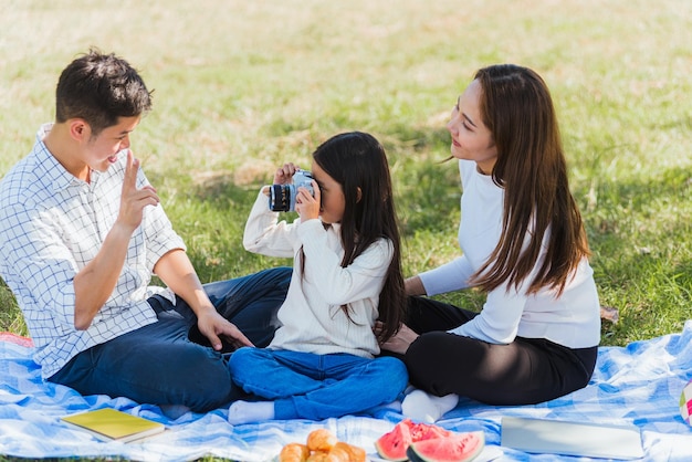 Felice giovane famiglia asiatica padre, madre e figlio bambina che si diverte e si gode un picnic all'aperto insieme seduti su una coperta da picnic che scatta foto con una fotocamera retrò nel parco giardino in una giornata di sole