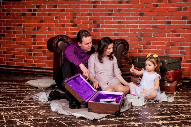 Felice giovane famiglia con una bambina di tre anni seduta in soffitta con vecchie valigie e giocando in modo carino. Bella madre incinta, padre e figlia a casa e giocano. Momenti d'atmosfera in famiglia del bambino'