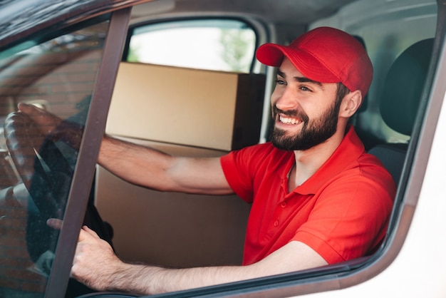 felice giovane fattorino in uniforme rossa sorridente e guida furgone con cassette dei pacchi