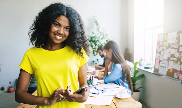 Felice giovane fiduciosa bella donna d'affari africana in abbigliamento casual intelligente è in posa su sfondo moderno team di affari. Collaboratori. Avvia squadra. Studenti insieme