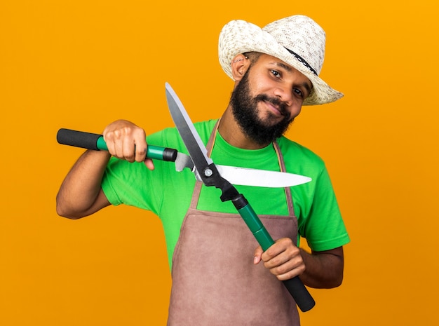 Felice giovane giardiniere afro-americano che indossa un cappello da giardinaggio che tiene le forbici isolate sulla parete arancione