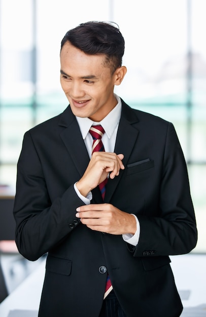 Felice giovane imprenditore maschio etnico asiatico in abito formale in piedi e regolare la manica del vestito in ufficio.