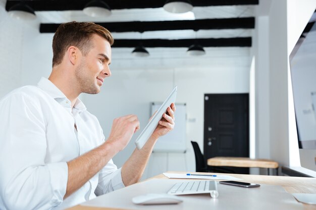 Felice giovane imprenditore seduto e utilizzando tablet in office