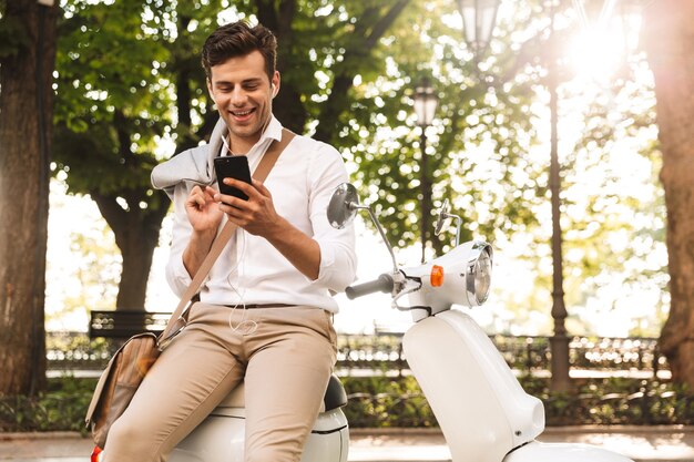 Felice giovane imprenditore seduto su una moto all'aperto, utilizzando il telefono cellulare