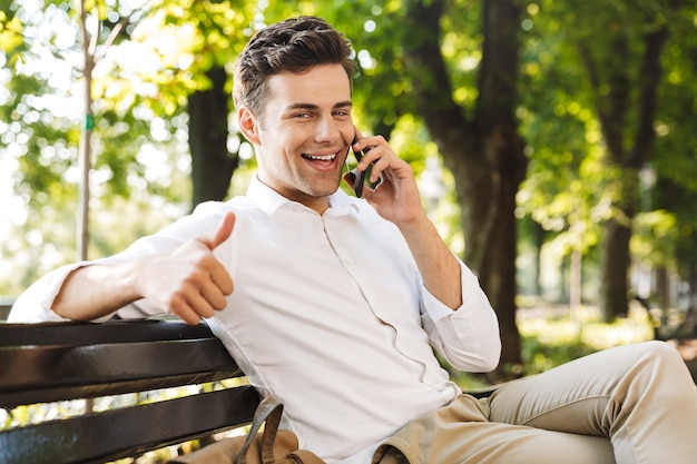 Felice giovane imprenditore seduto su una panchina all'aperto, parlando al telefono cellulare